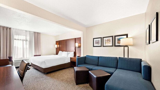A hotel room with a large bed, a sofa, a desk, and a chair. There are framed pictures on the wall and a lamp on the desk.