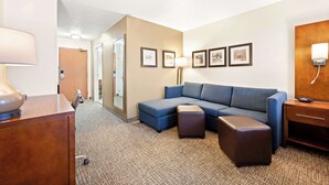 A hotel room with a sectional sofa, a desk with a lamp, a bedside table, and framed pictures on the wall.