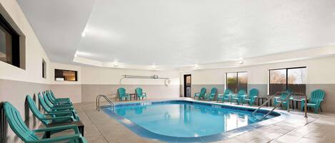 A large indoor swimming pool with blue tiles, surrounded by turquoise chairs and tables, and a tiled floor.
