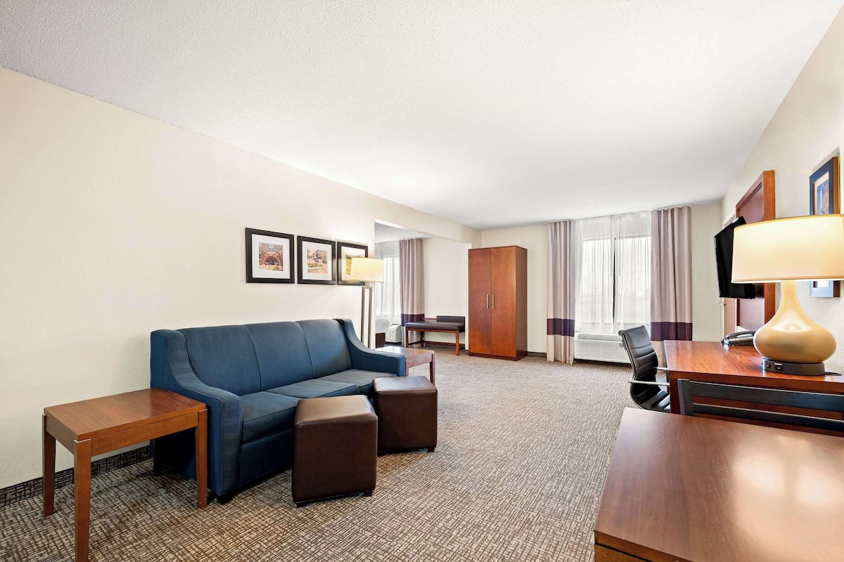 A hotel room with a blue sofa, a wooden table, a wooden cabinet, and a desk with a lamp.