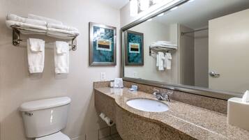A modern bathroom with a white toilet, a granite sink, and a large mirror. There are towels on a rack and a framed artwork on the wall.