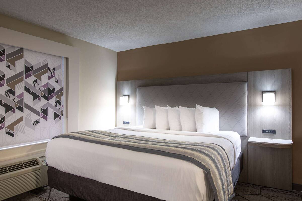 Une chambre d’hôtel moderne dotée d’un grand lit, d’une tête de lit avec éclairage intégré, d’un climatiseur mural et d’une décoration murale aux motifs géométriques.