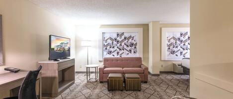 A hotel room with a desk, a television, a sofa, ottomans, and patterned artwork on the walls.
