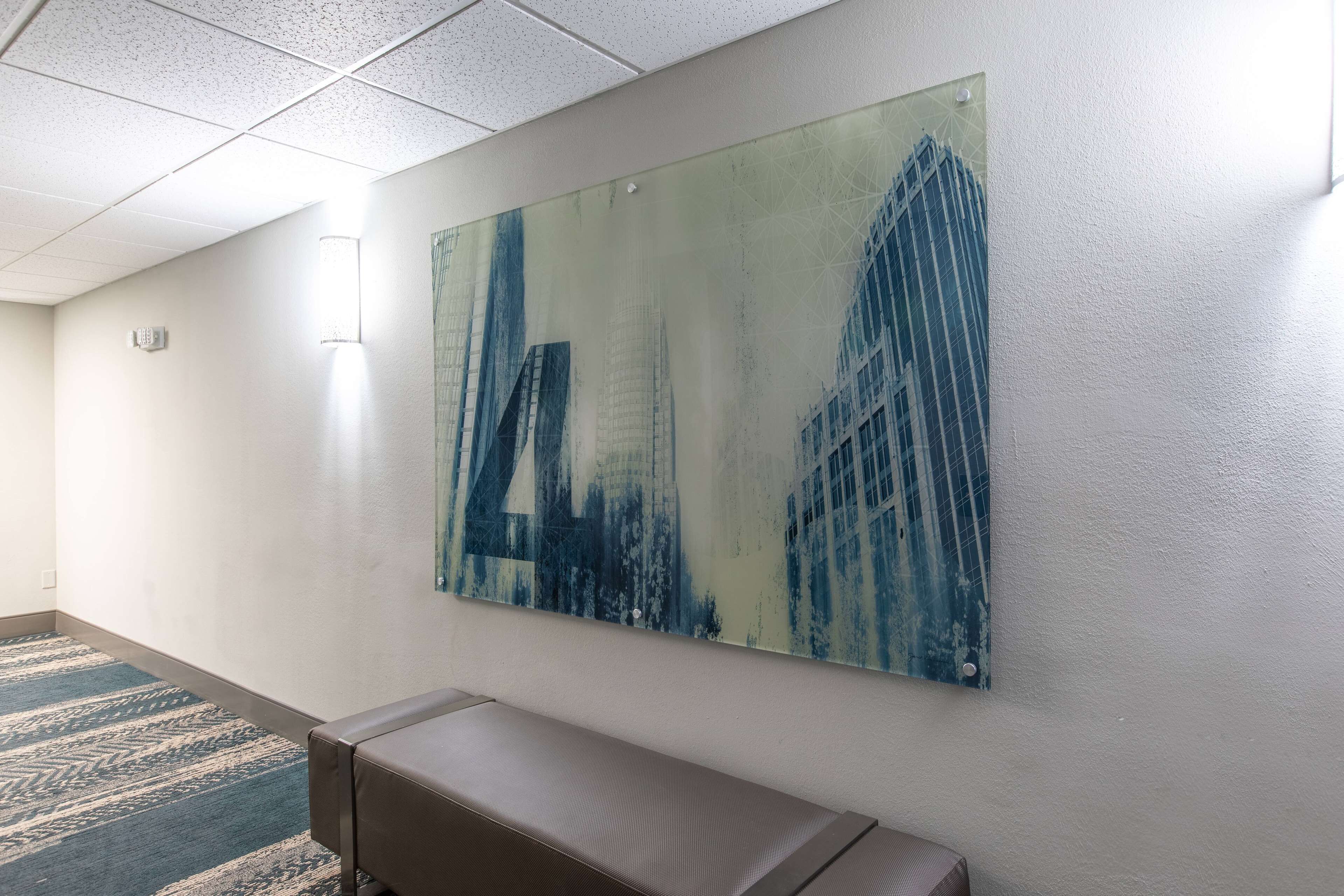 A hallway with a large abstract painting of buildings on the wall, a bench, and patterned carpet.