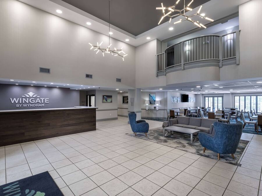A spacious lobby with a reception desk labeled "Wingate by Wyndham," modern seating, and a high ceiling with recessed lighting.