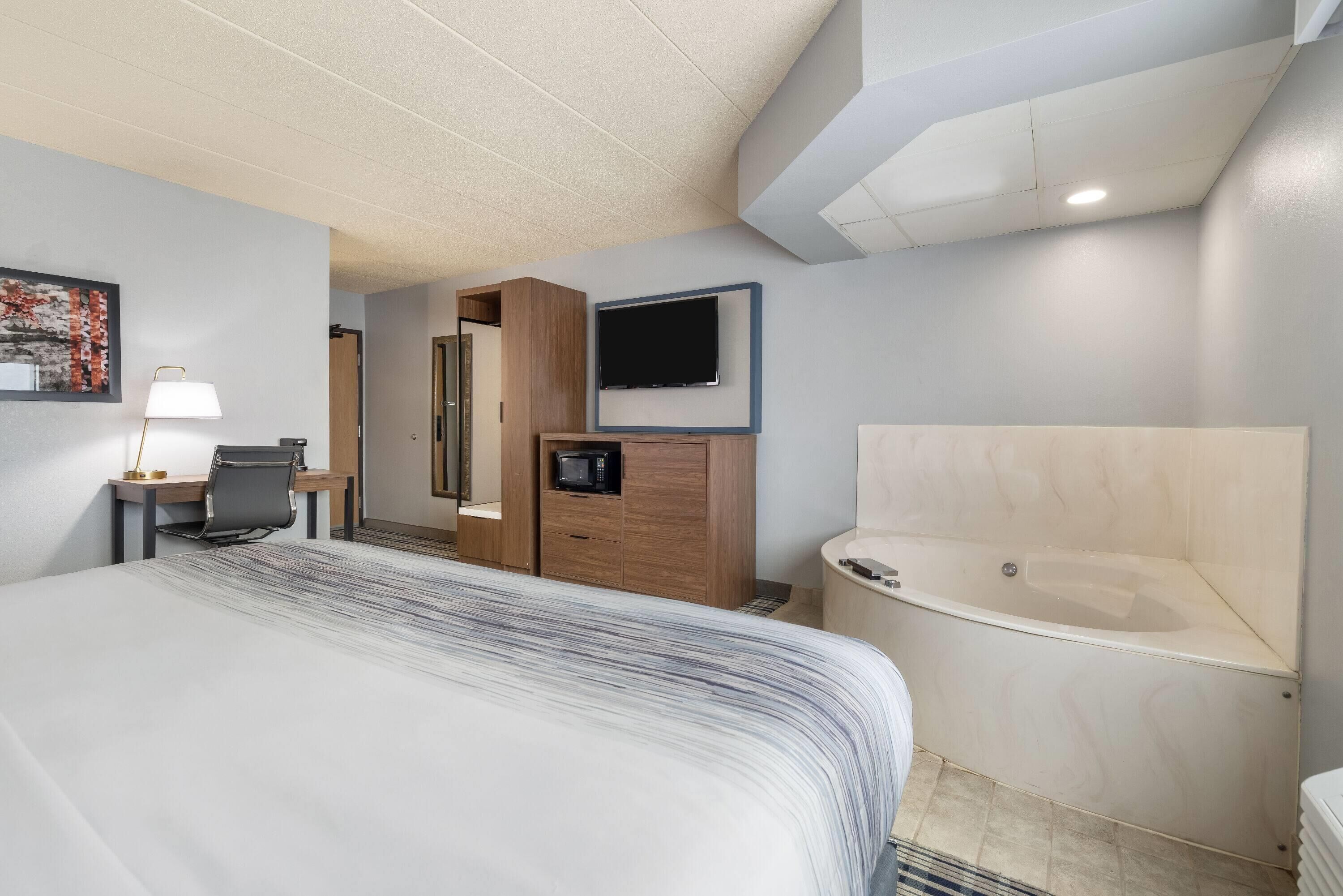 Een hotelkamer met een groot bed, een bureau met stoel, een aan de muur bevestigde televisie, een houten garderobe en een bad.