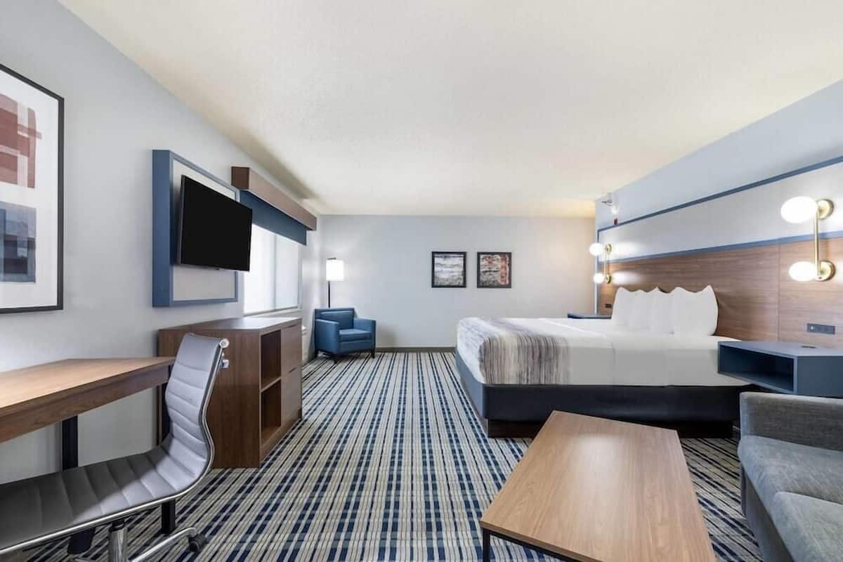 A hotel room with a bed, a desk, a chair, a TV, and a wall-mounted light fixture.