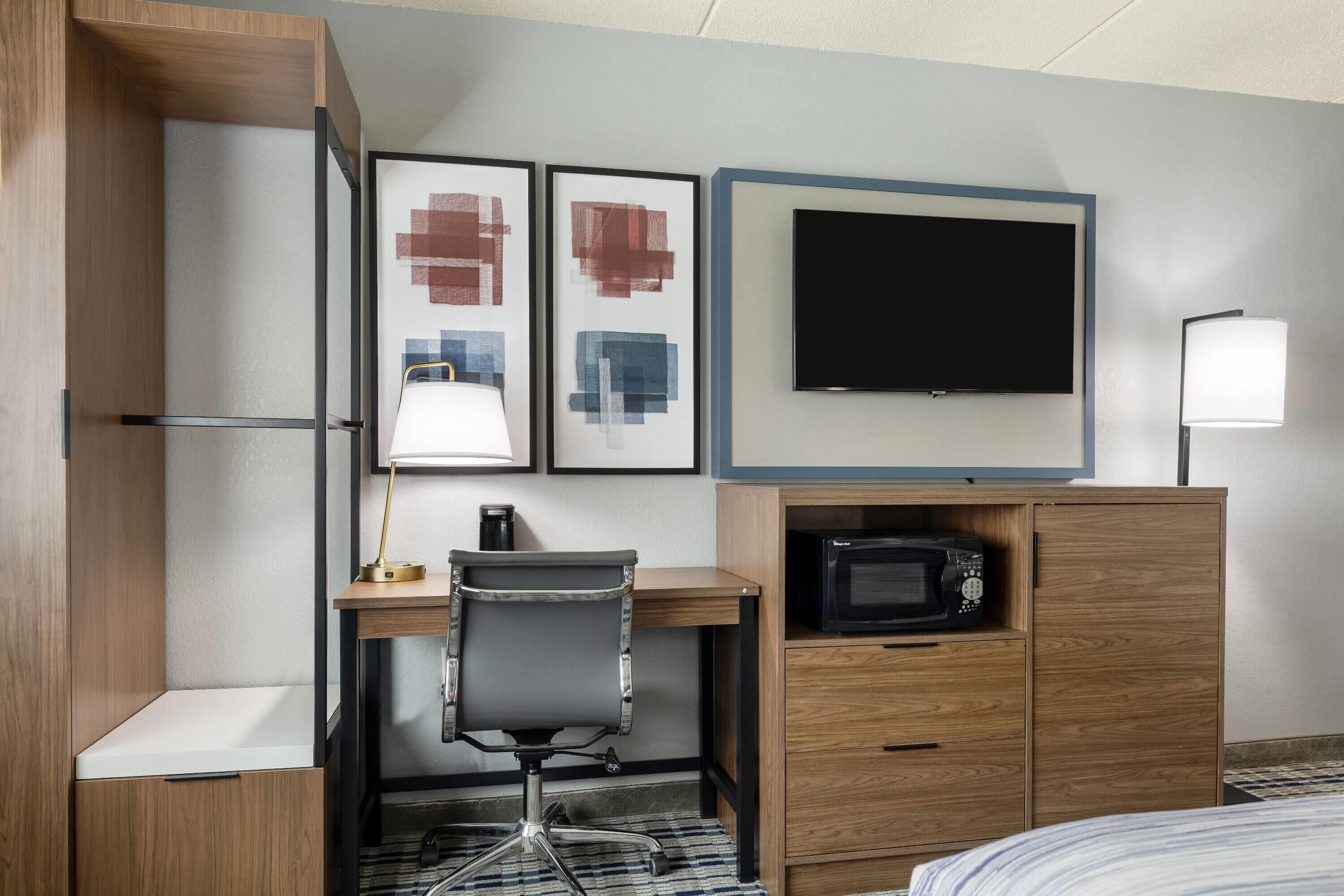 A hotel room with a wooden desk, a chair, a flat-screen TV, a microwave, a bedside lamp, and two abstract paintings.