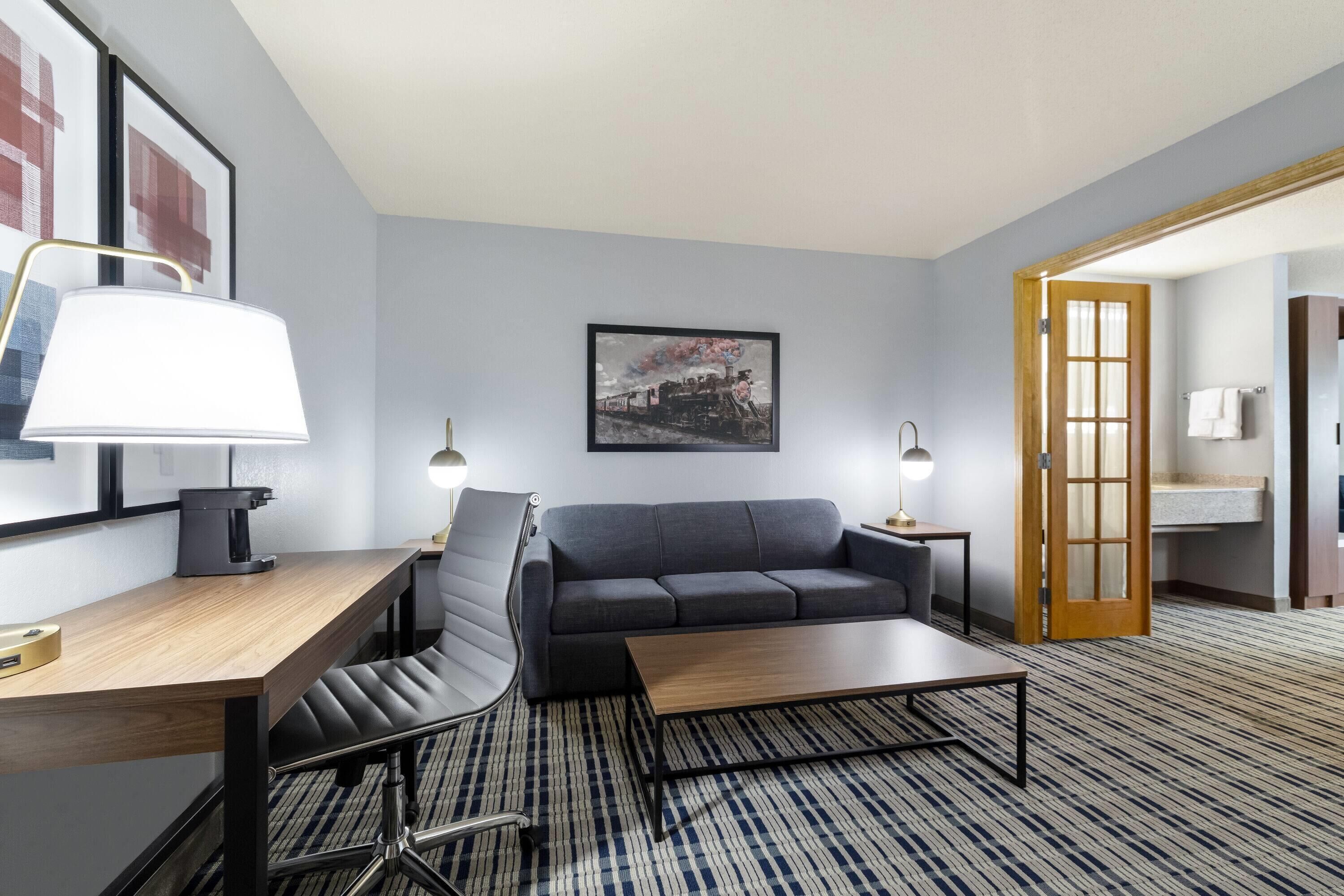 Quarto de hotel moderno com uma escrivaninha de madeira, uma poltrona de couro preto, um sofá cinzento e uma mesa de centro de madeira, além de um tapete estampado.
