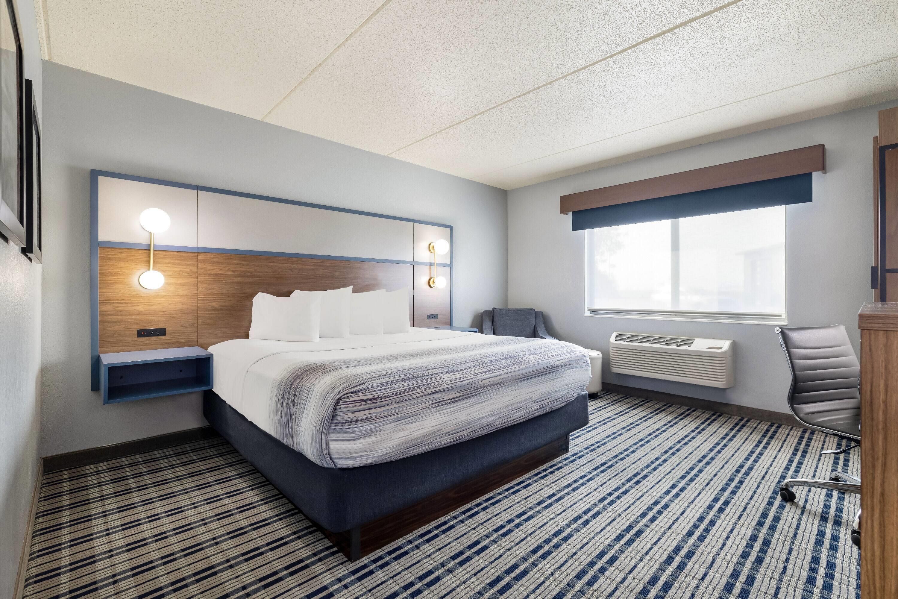 A hotel room with a large bed, bedside tables, a chair, a window with blinds, and a wall-mounted air conditioner.