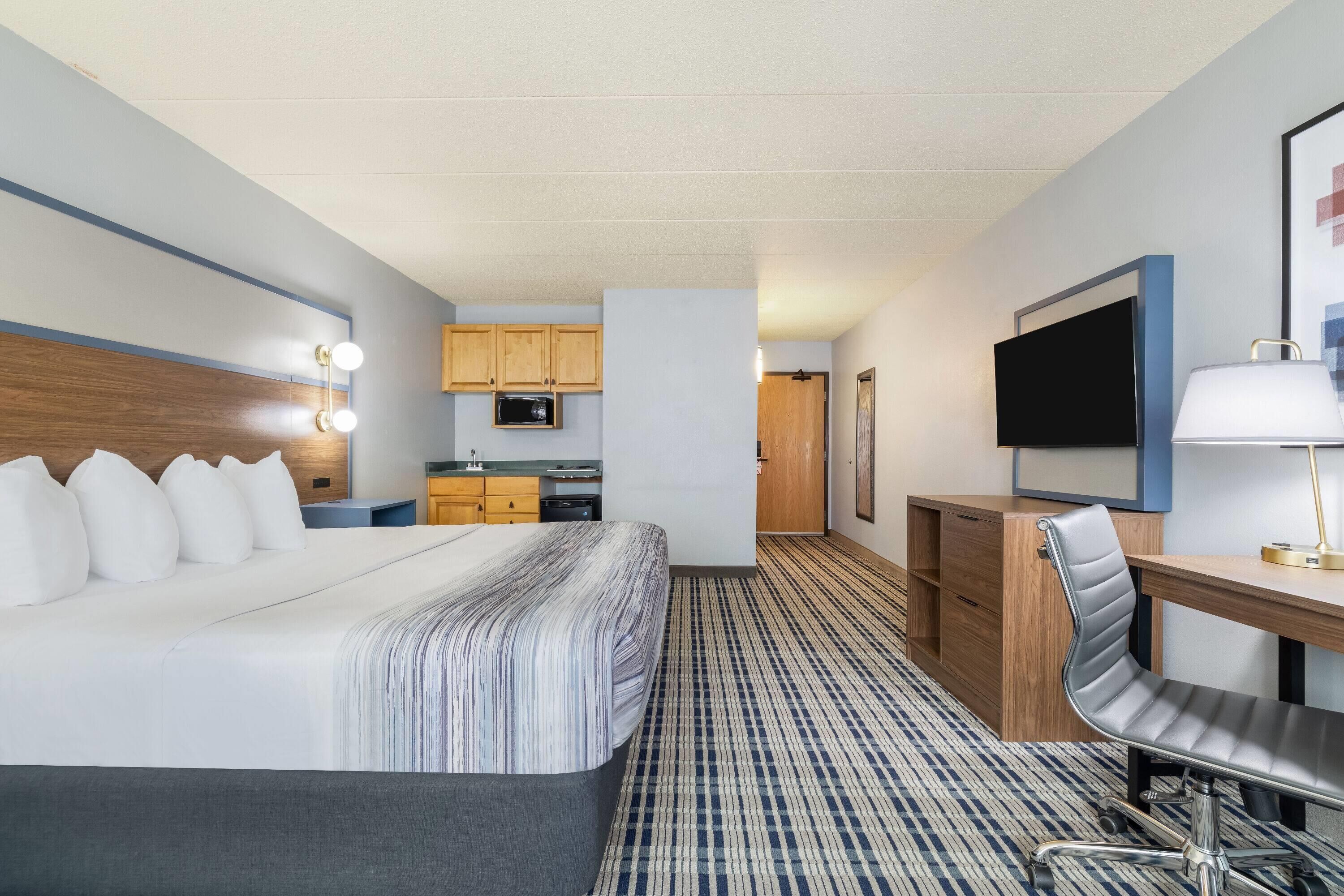 A hotel room with a large bed, a desk with a chair, a television, and a kitchenette.