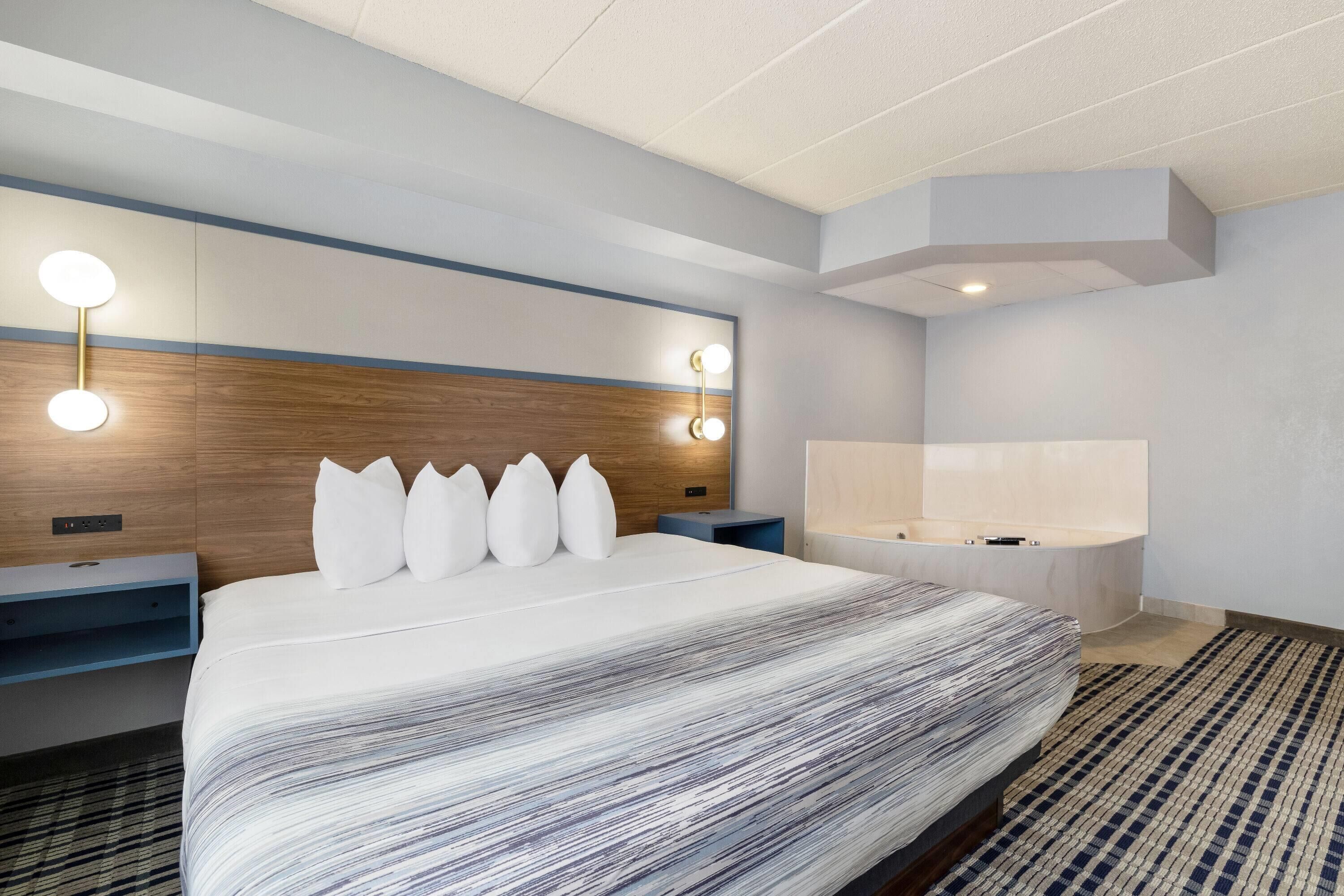 A modern hotel room with a large bed, bedside tables, a vanity with a mirror, and a built-in bathtub.