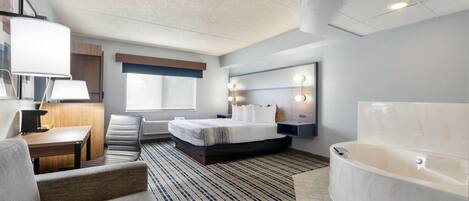 Habitación de hotel con cama, escritorio, silla, ventana grande, baño con bañera y tocador con luces.
