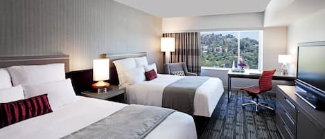 Habitación de hotel con dos camas, un escritorio, una silla, un televisor y ventana con vista a la ciudad.