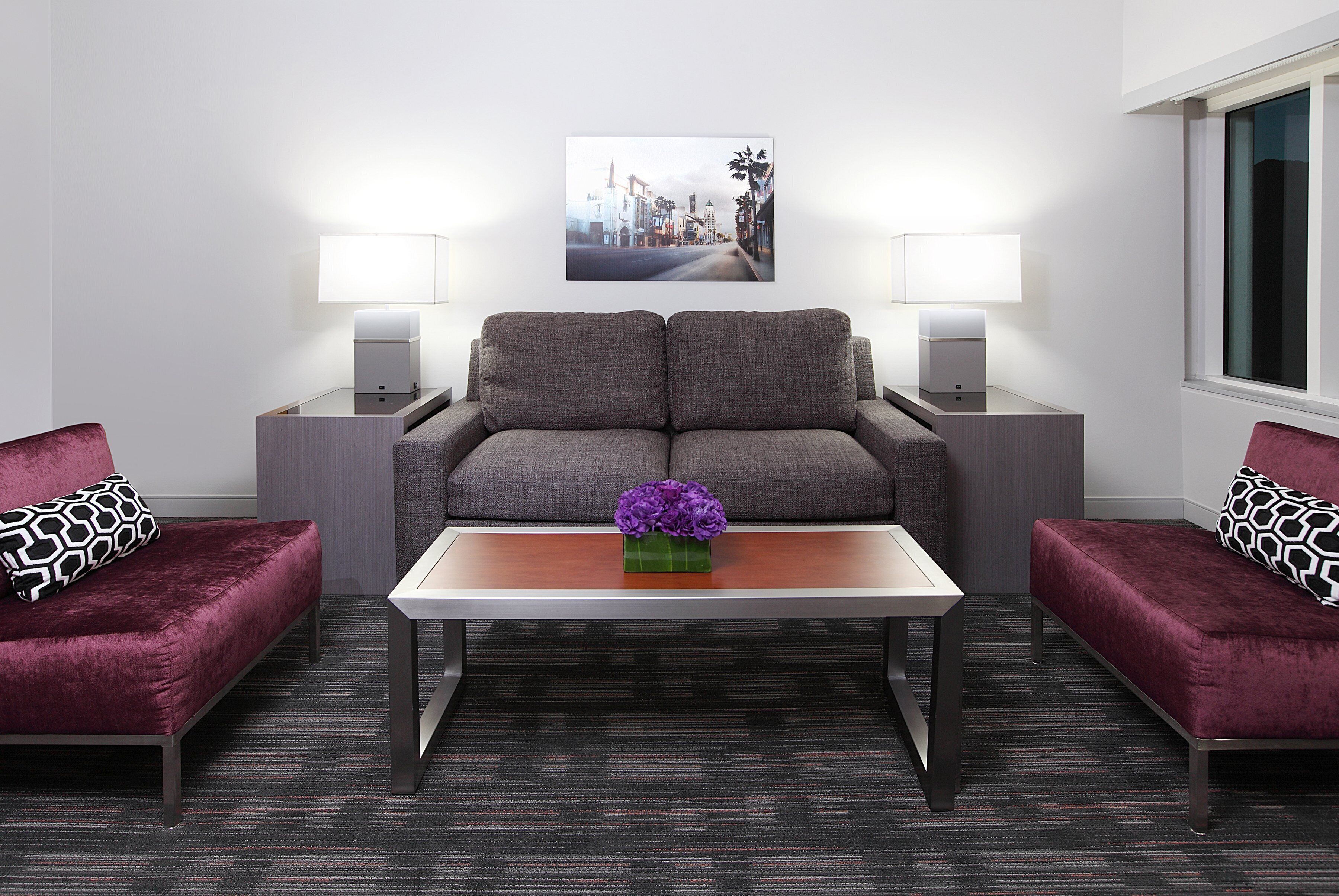 A modern hotel room with a grey sofa set, a wooden coffee table with a purple flower vase, and two bedside tables with lamps.