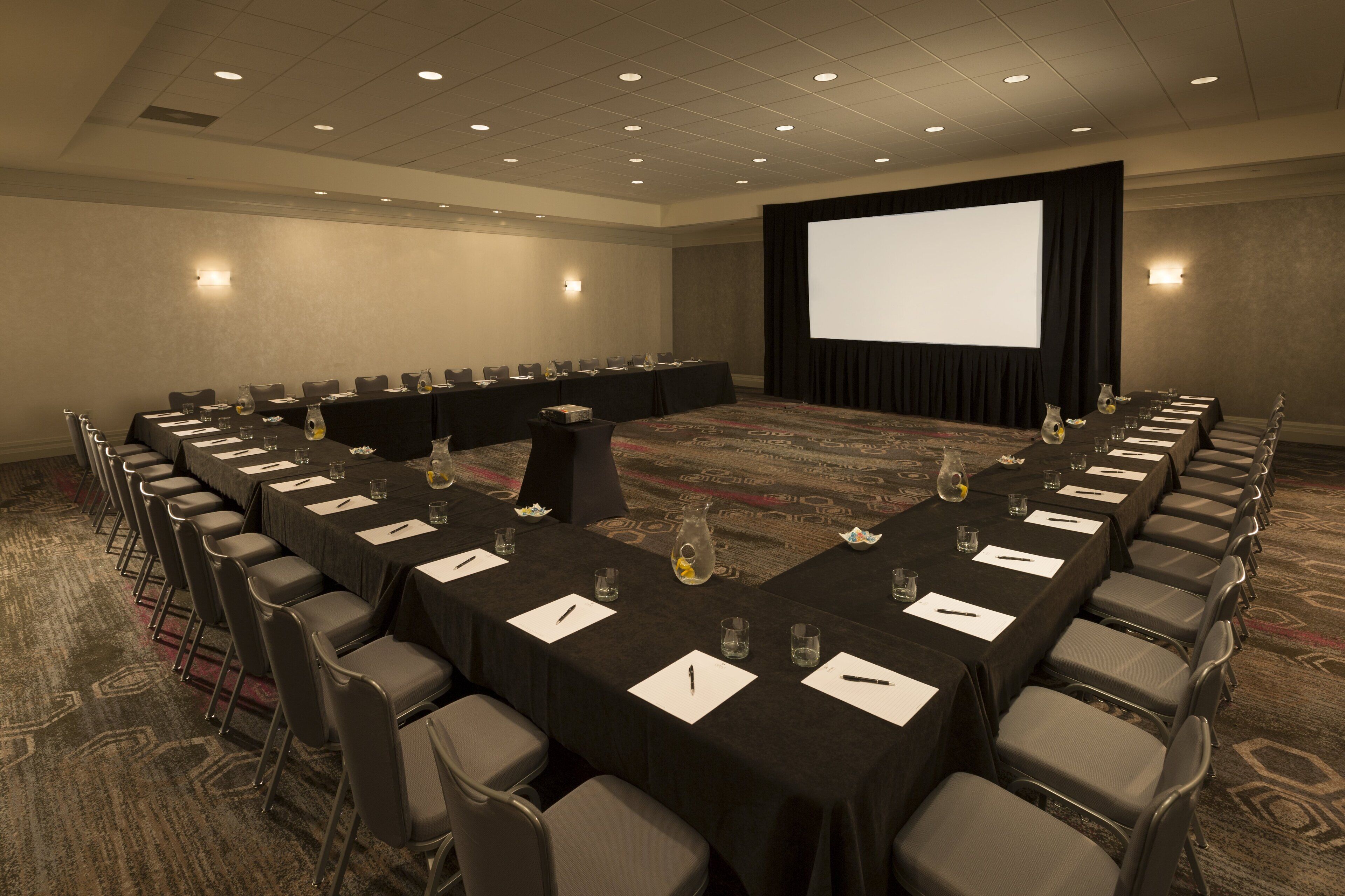 A conference room with a long table, multiple chairs, a projector screen, and a carpeted floor.