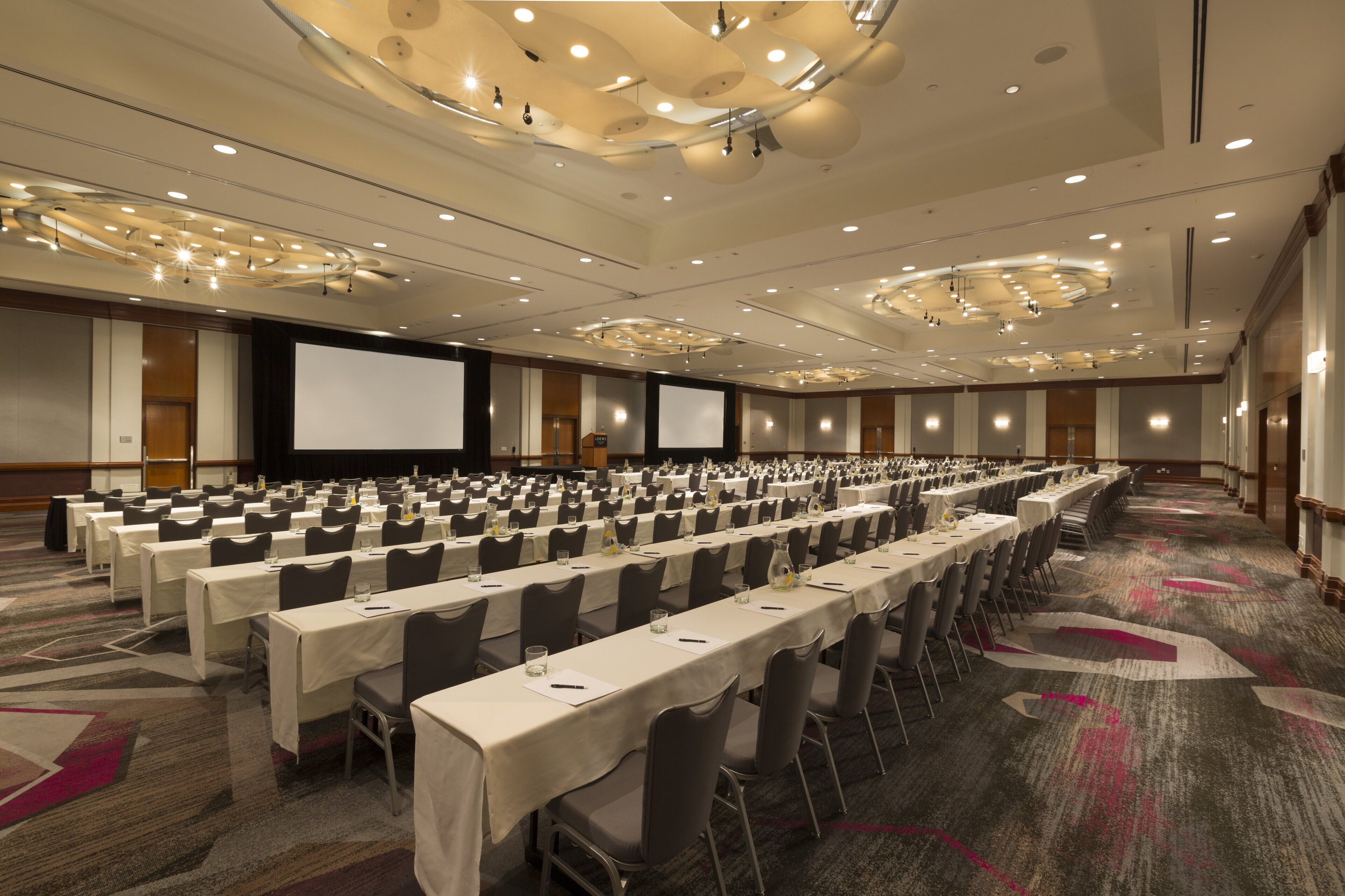 A large conference room with rows of tables and chairs, a projector screen, and a carpeted floor.