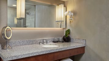 A bathroom with a granite countertop, a white sink, a mirror with a gold frame, a black vase with a plant, and a wall-mounted light fixture.