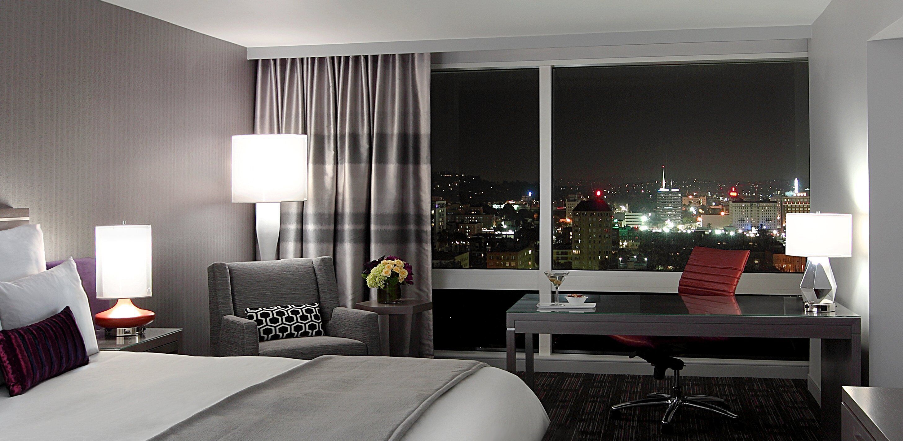 Une chambre d’hôtel avec un lit, un bureau, une chaise, une table et une vue sur le paysage urbain la nuit.