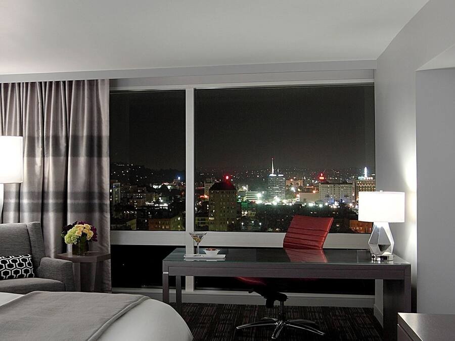 Quarto de hotel com cama, escrivaninha, cadeira, televisão e janela com vista para a cidade.