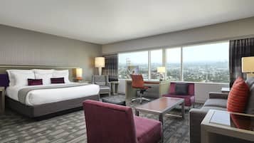 Una habitación de hotel con una cama grande, un escritorio, una silla, un sofá y vistas de la ciudad a través de amplios ventanales.