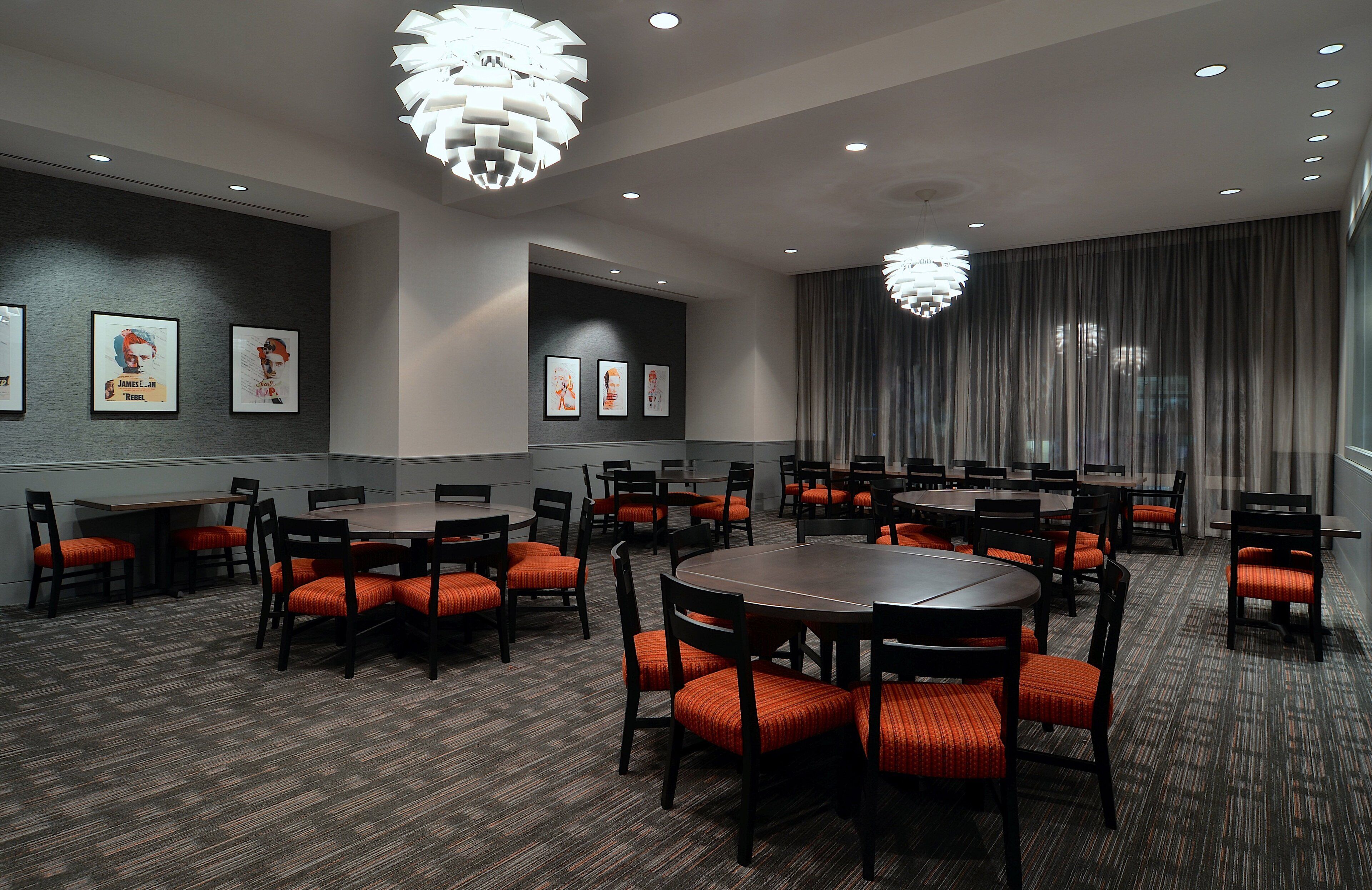 A conference room with round tables and orange chairs, featuring ceiling lights and framed artwork on the walls.