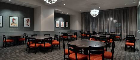 A conference room with round tables and orange chairs, featuring ceiling lights and framed artwork on the walls.