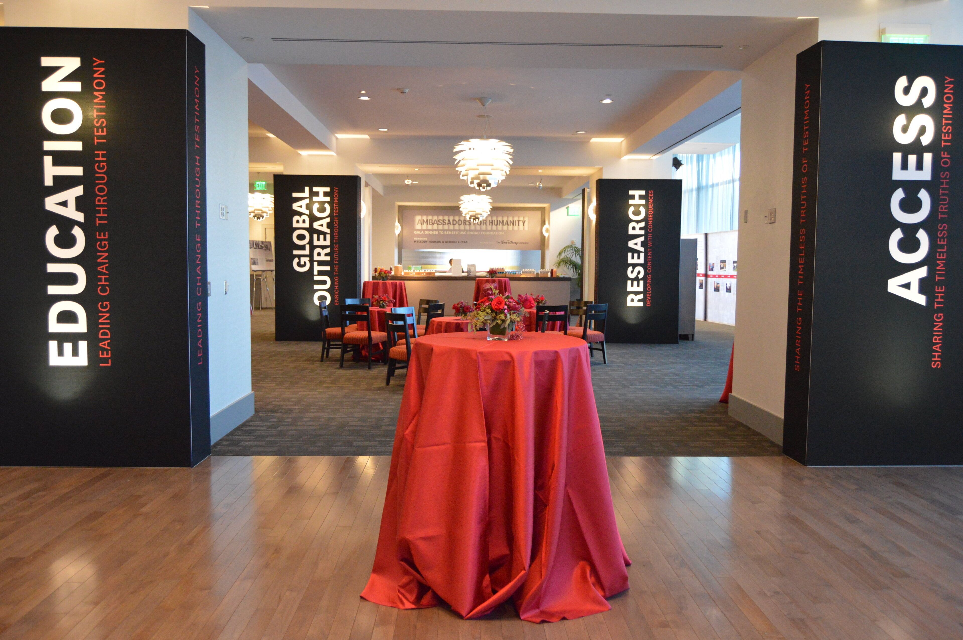 A modern hotel lobby with a central round table covered in a red tablecloth, flanked by black pillars with white text.