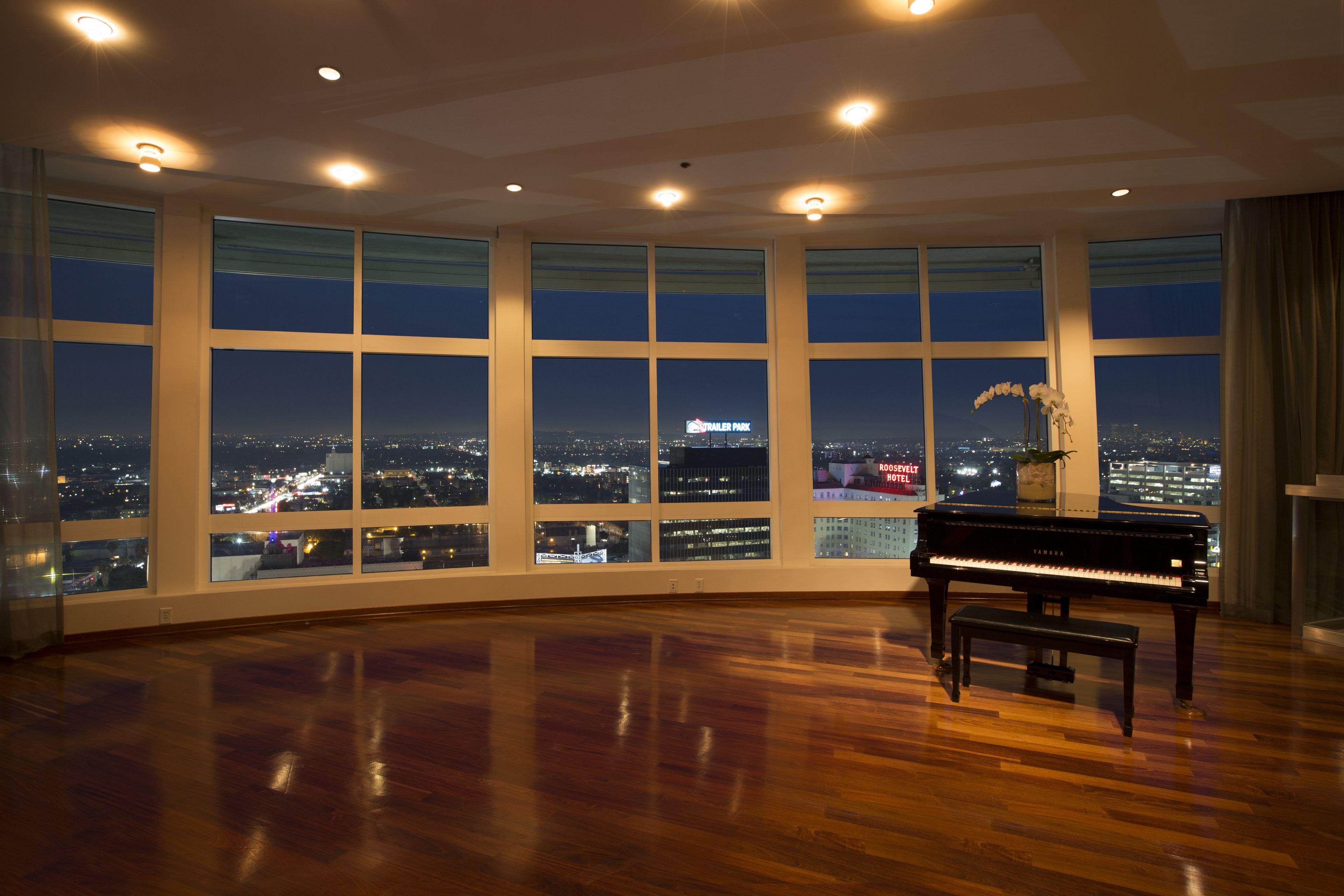 A spacious room with a grand piano, large windows offering a city view, and a modern ceiling design.