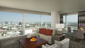 Une chambre d’hôtel moderne avec vue sur la ville, comprenant un canapé, une table basse avec des fruits, une lampe et un coin repas.