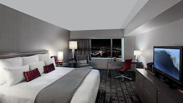 A modern hotel room with a large bed, a desk, a TV, and a view of the city at night.