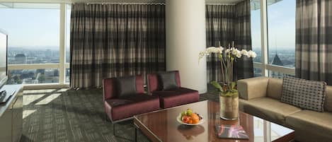 Habitación de hotel moderna con un ventanal amplio que ofrece vistas a la ciudad, un sofá, una mesita de centro con fruta y un jarrón con flores.