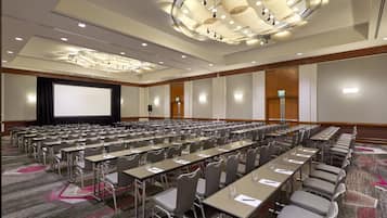 Une grande salle de conférence avec de nombreuses tables et chaises disposées en rangées, un écran de projection et un lustre moderne.