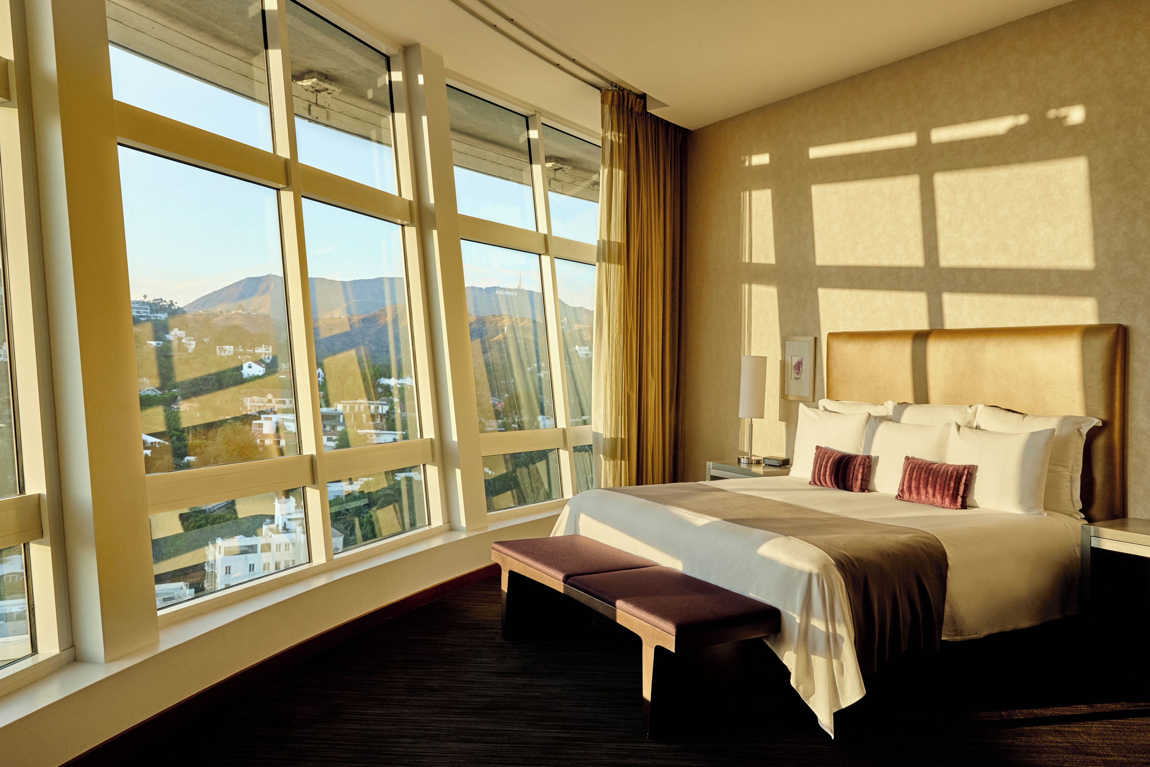 A hotel room with a large window, a bed with pillows, a bedside table, and a bench.