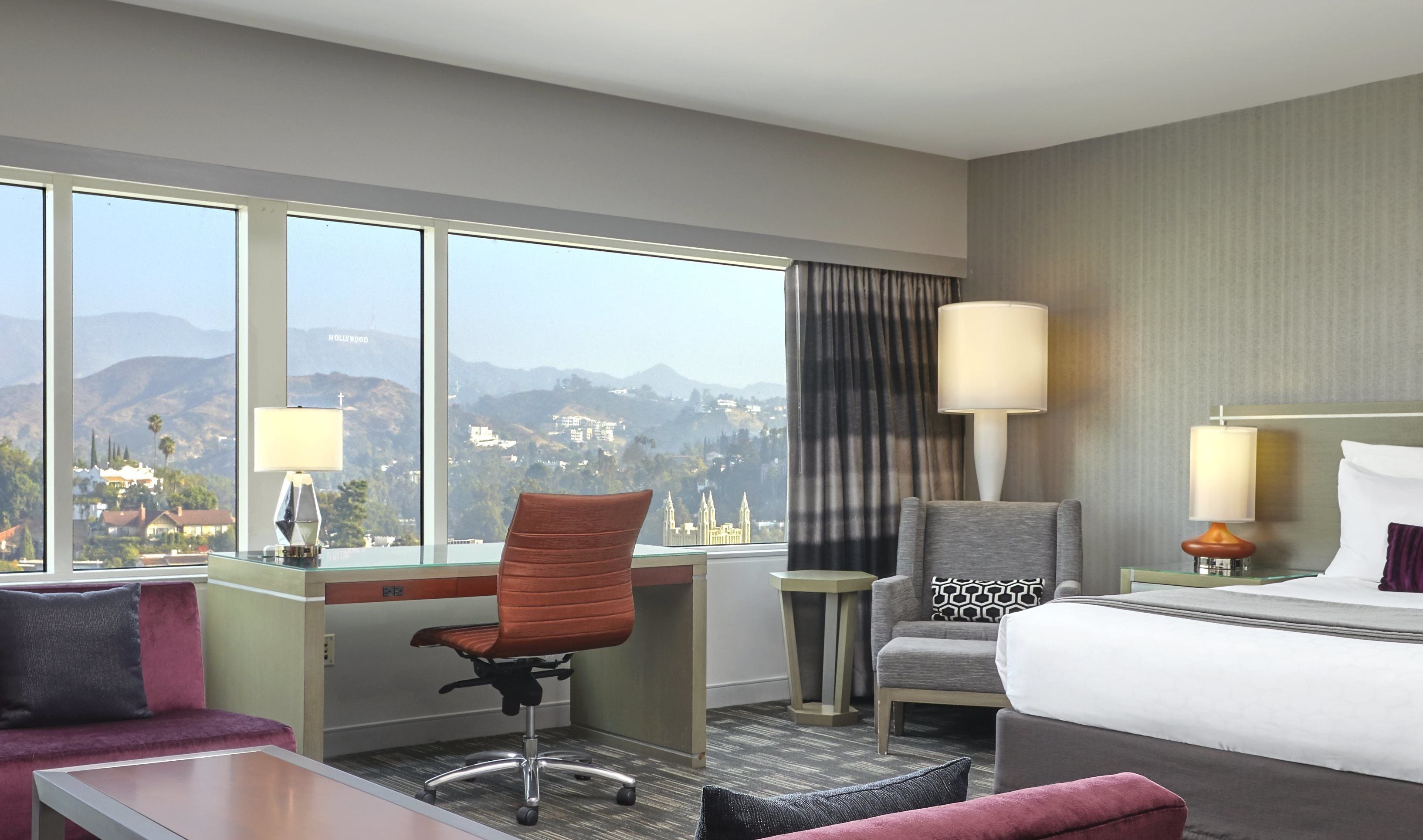 A hotel room with a desk, office chair, and a bed. There is a view of mountains and a cityscape through large windows.
