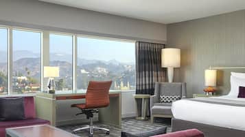 A hotel room with a desk, office chair, and a bed. There is a view of mountains and a cityscape through large windows.