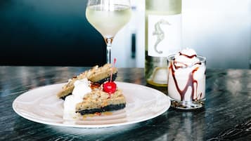 Un plato de postre con una porción de pastel, una copa de vino blanco y un vaso pequeño de postre con salsa de chocolate y crema batida.