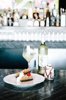 Un plato de postre con una porción de pastel, una copa de vino blanco y un vaso pequeño de postre con salsa de chocolate y crema batida.