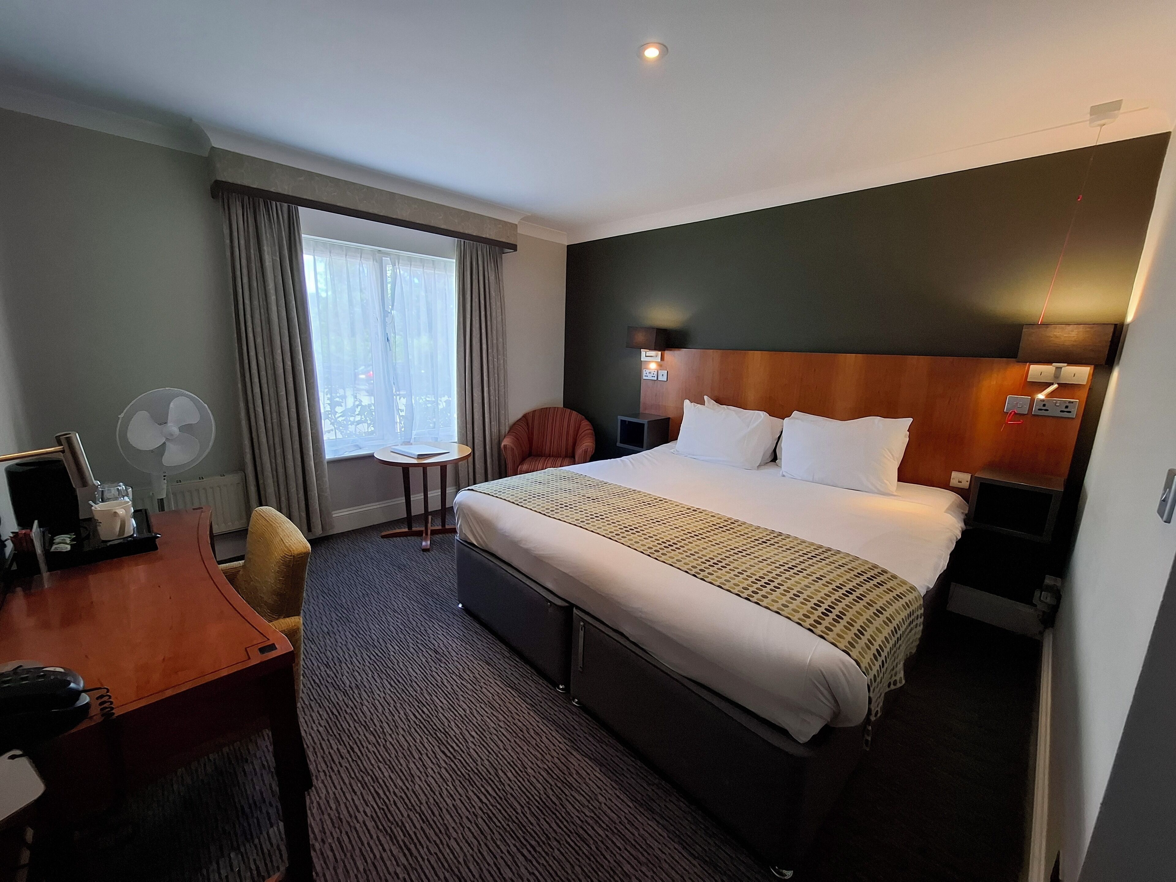 Habitación de hotel con una cama grande, un escritorio, una silla, una mesita y un ventanal con cortinas.