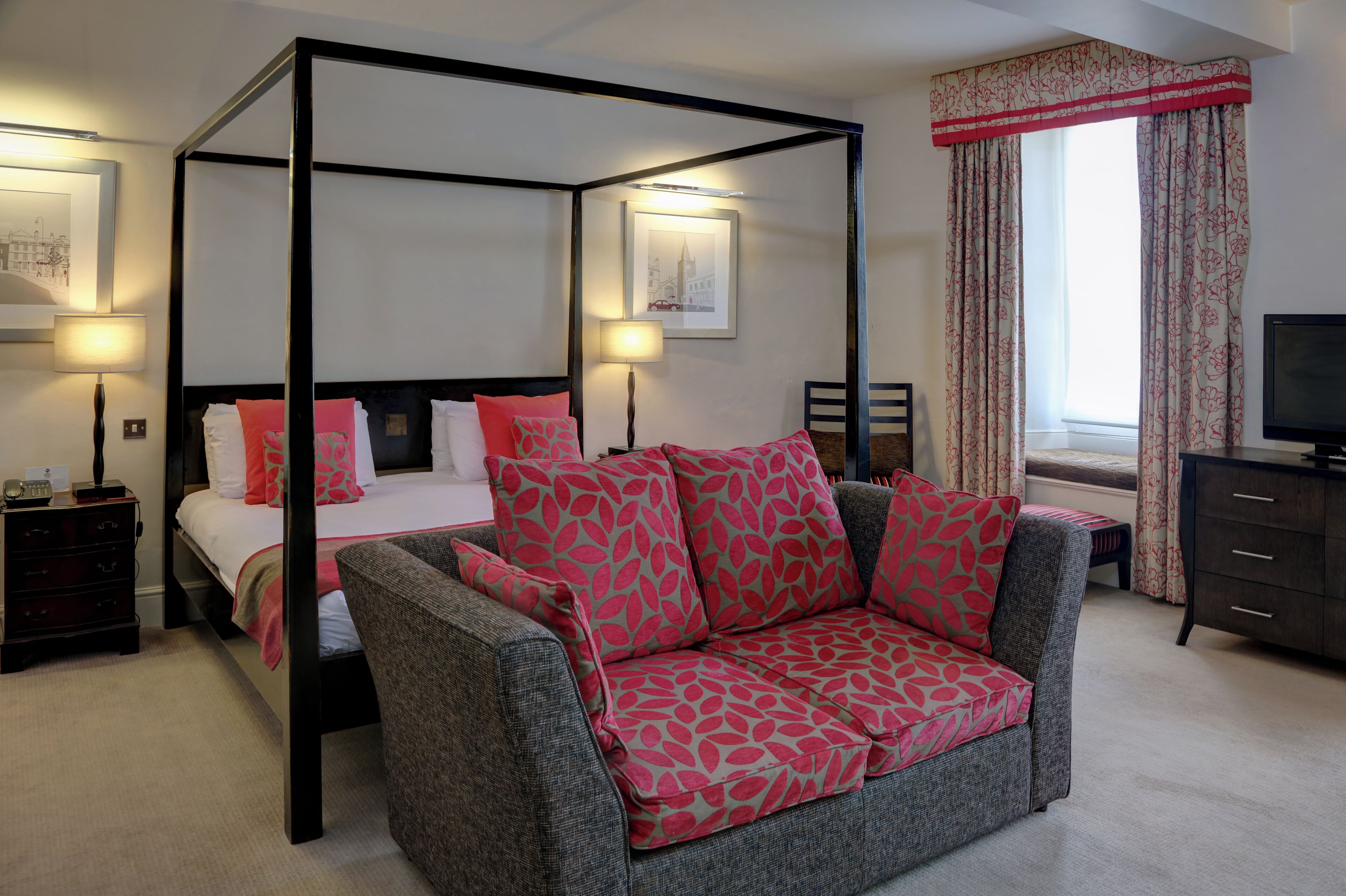 A four-poster bed with a white and red bedspread, a grey sofa with red leaf-patterned cushions, a wooden nightstand with a lamp, and a television on a dark wooden cabinet.