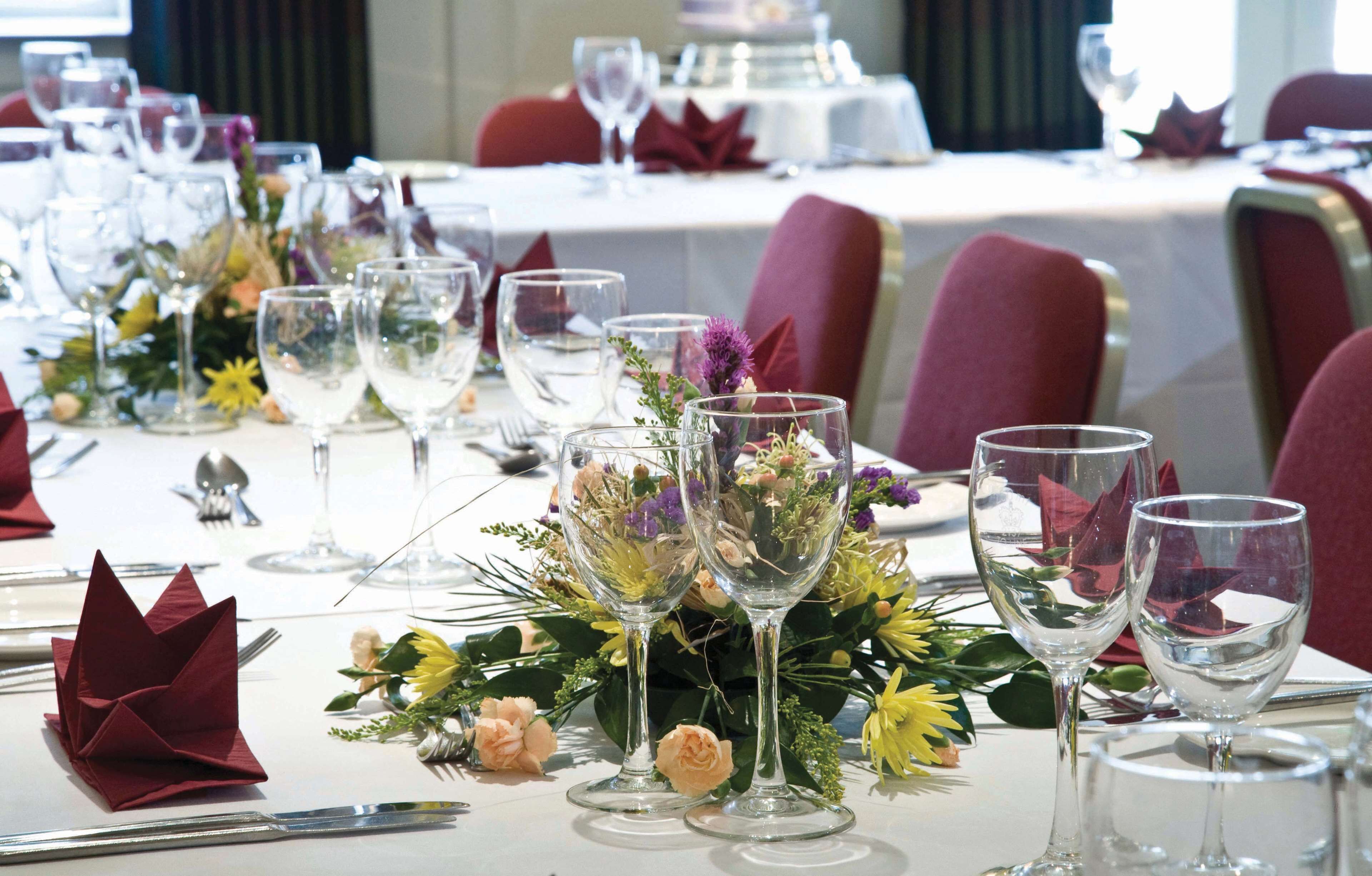 Una mesa de banquete dispuesta con cubertería de vidrio, servilletas rojas y centros de mesa florales.