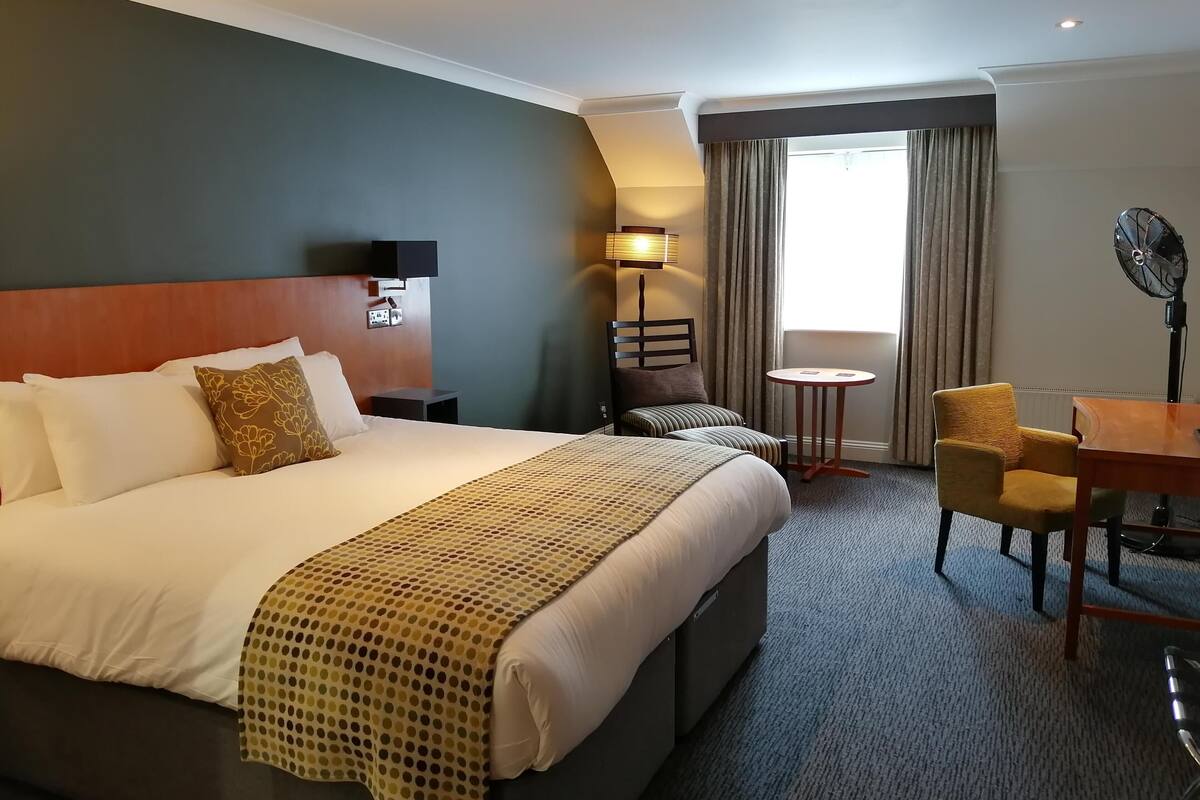A hotel room with a large bed, a small table, a chair, a desk, a lamp, and a fan.