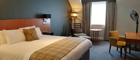 Habitación de hotel con una cama grande, una mesita pequeña, una silla, un escritorio, una lámpara y un ventilador.