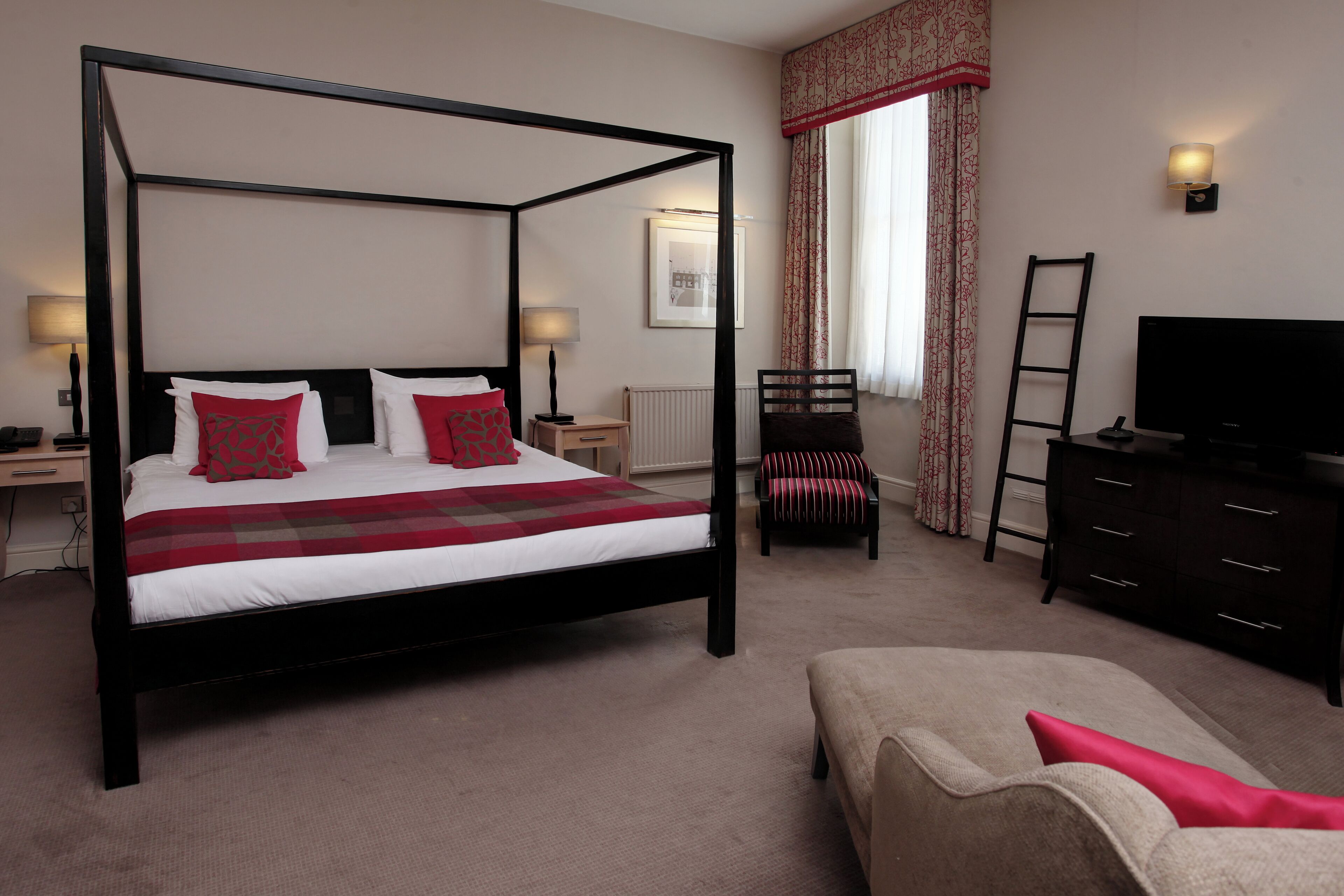A four-poster bed with red and white bedding, a television, a desk with a lamp, and a chair.