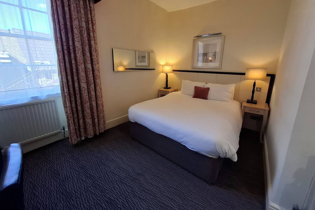 A hotel room with a large bed, two bedside tables with lamps, a window with curtains, and a framed picture on the wall.