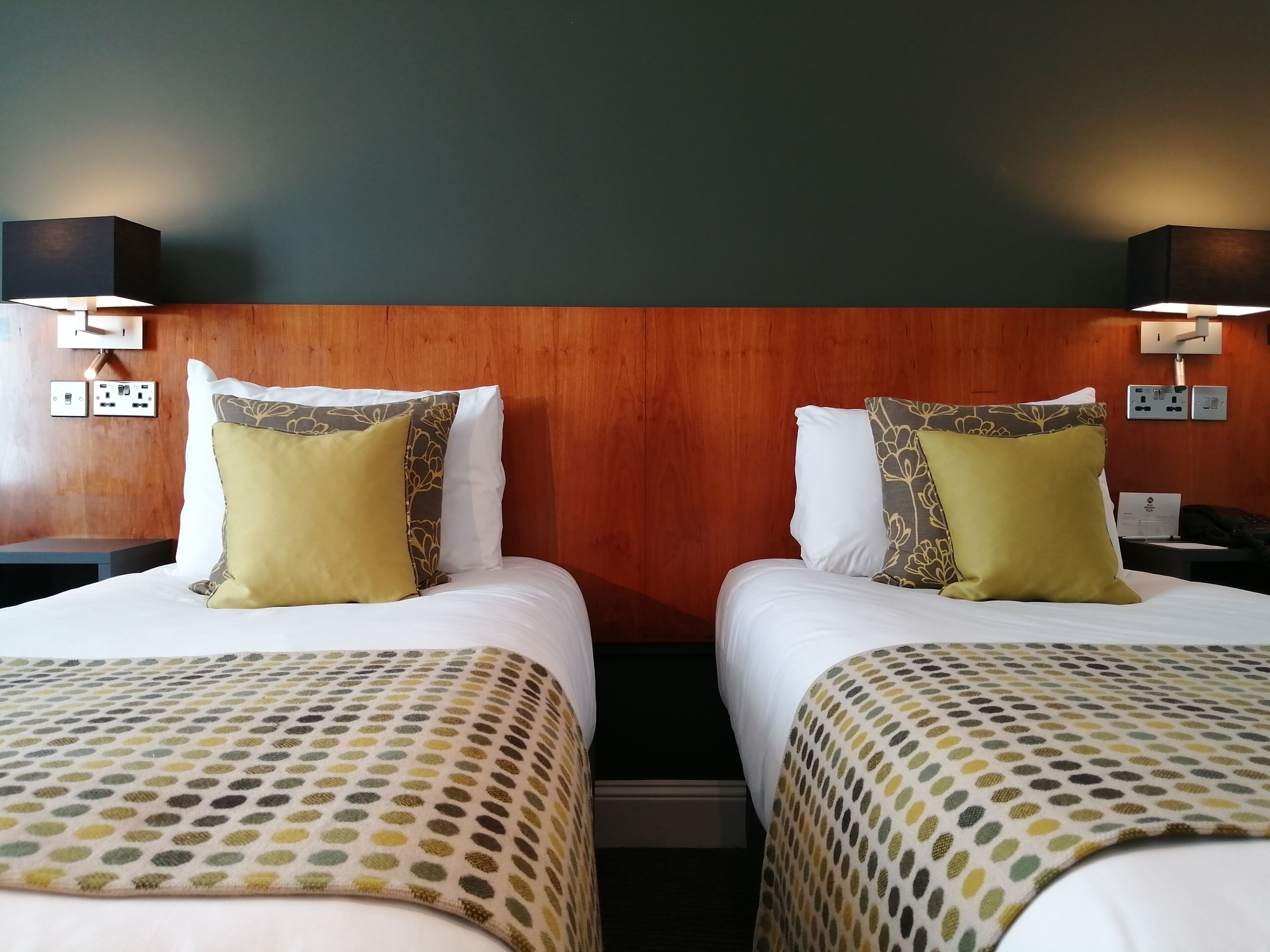 A hotel room with two beds, each with a patterned bedspread, a wooden headboard, and wall-mounted lamps.