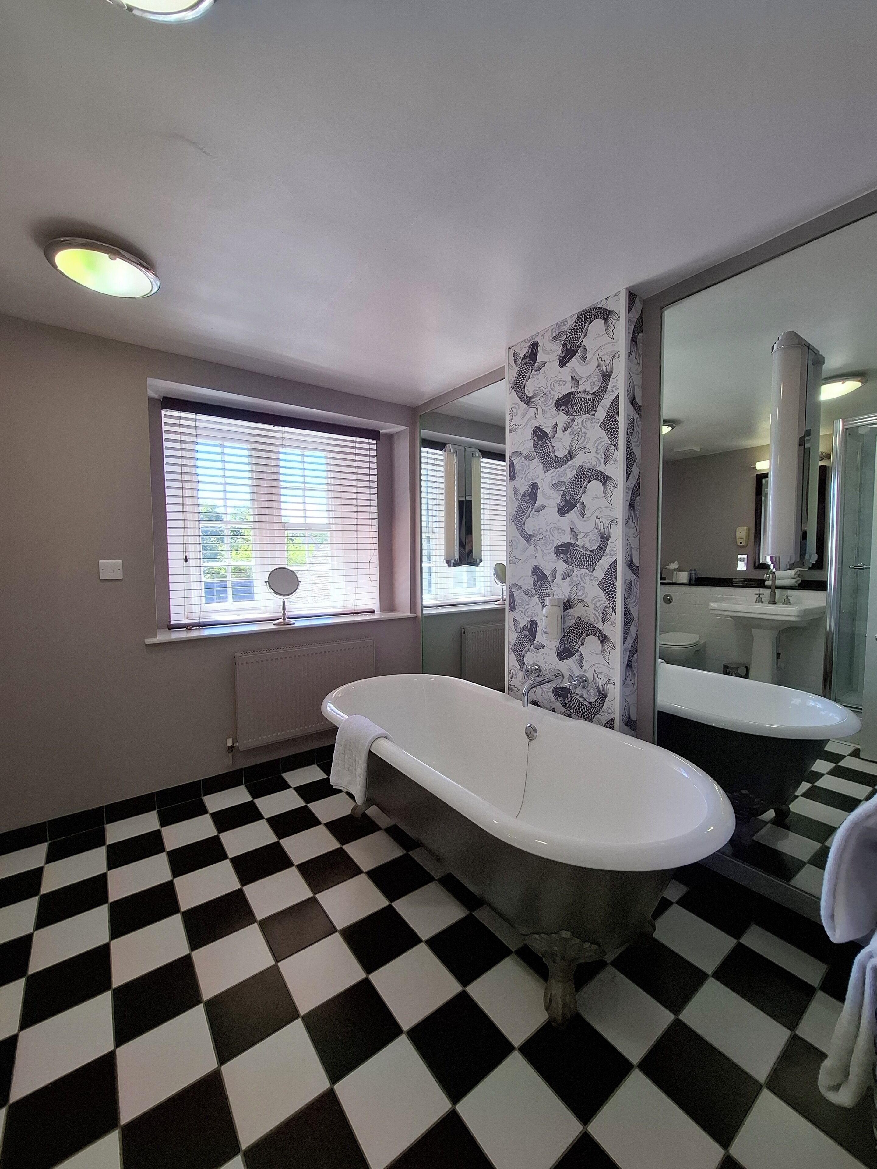 Een badkamer met een geruite vloer, een witte badkuip, een grote spiegel en een raam met jaloezieën.