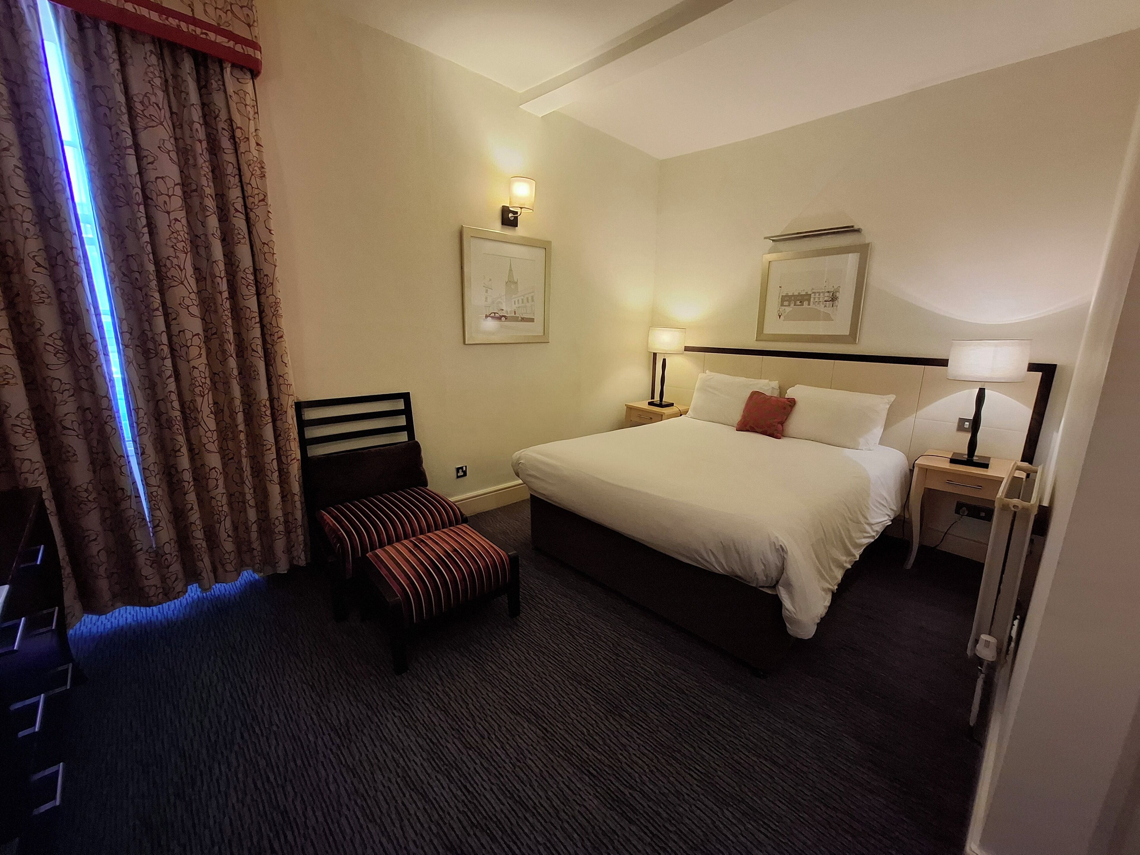 Une chambre d’hôtel avec un lit, deux tables de chevet avec des lampes, un petit banc et deux tableaux encadrés au mur.