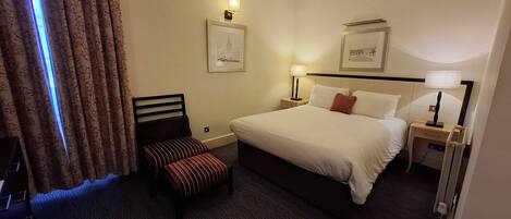 Une chambre d’hôtel avec un lit, deux tables de chevet avec des lampes, un petit banc et deux tableaux encadrés au mur.