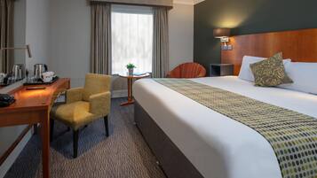 Habitación de hotel con una cama grande, un escritorio con silla, una mesita con planta y una ventana con cortinas.